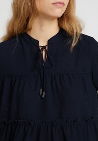 Femme portant un blouse bleu marine avec un col rond noué avec un nœud et des perles décoratives sur les cordons.