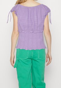 Top smockato viola con maniche corte e nastri arricciati, caratterizzato da un orlo arricciato. Abbinato a pantaloni cargo verdi. Texture liscia.