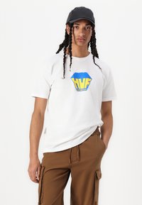 Hvit t-skjorte med en blå og gul "HUF" logo; sammen med brune cargo-bukser; modellen har på seg en svart caps med jevn tekstur.