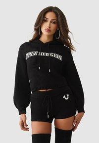 Schwarzer, gerippter, verkürzter Hoodie mit "TRUE RELIGION"-Logo, kombiniert mit schwarzen Shorts mit Kordelzug und Emblem an der Seite.