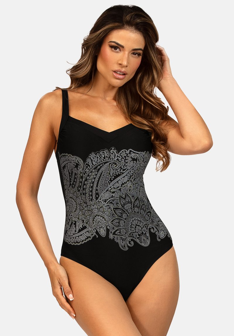 Kobieta w czarnym jednoczęściowym kostiumie kąpielowym z szerokimi ramiączkami i szczegółowym srebrnym wzorem paisley na torsie, pozująca z jedną ręką dotykającą włosów.