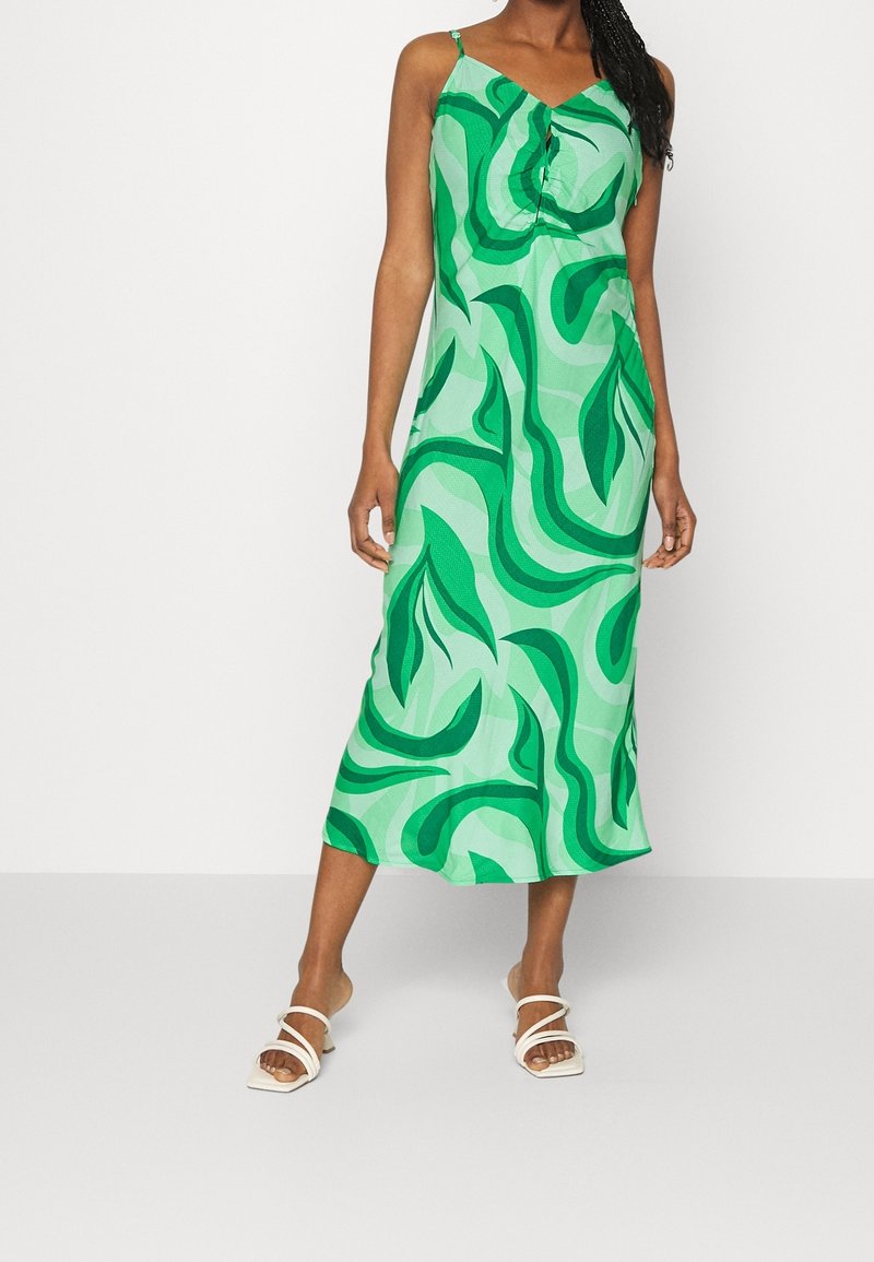 Femme portant une robe midi verte à motifs sans manches avec des bretelles spaghetti et des sandales blanches à talons, debout devant un fond uni.
