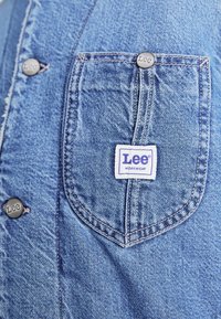 Denim overhemd in een lichtblauwe kleur, met een knoopsluiting aan de voorkant en een borstzak met een witte Lee® WORKWEAR-label en metalen knopen.