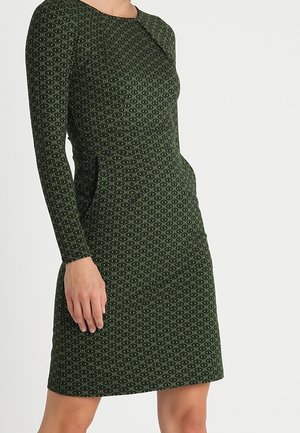 Kobieta w dopasowanej, zielonej sukience z długim rękawem z małym powtarzającym się geometrycznym wzorem, stojąca na jednolitym tle.