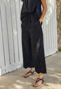 Schwarze, kurze Hosen mit bestickten Details und Ausschnitten, kombiniert mit braunen, schlichten Sandalen. Das Model steht vor einem rustikalen Holz-Hintergrund.
