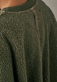 Grøn fleecejakke med en tekstureret, totteoverflade og en ribbet krave. Har en afslappet silhuet og en syning detalje langs ryggen.