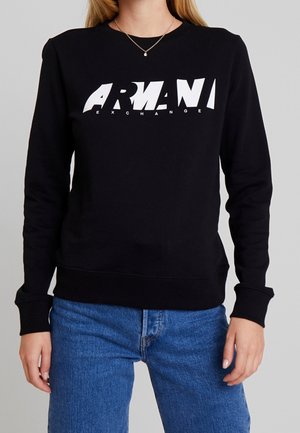 Femme portant un sweat-shirt noir Armani Exchange avec logo blanc et un jean bleu, fond gris uni.