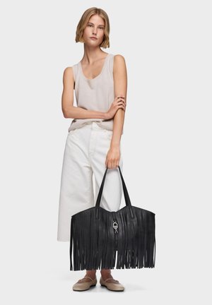 Mujer con top sin mangas beige y pantalones blancos de pierna ancha sosteniendo un gran bolso de cuero negro con flecos y cierre plateado.