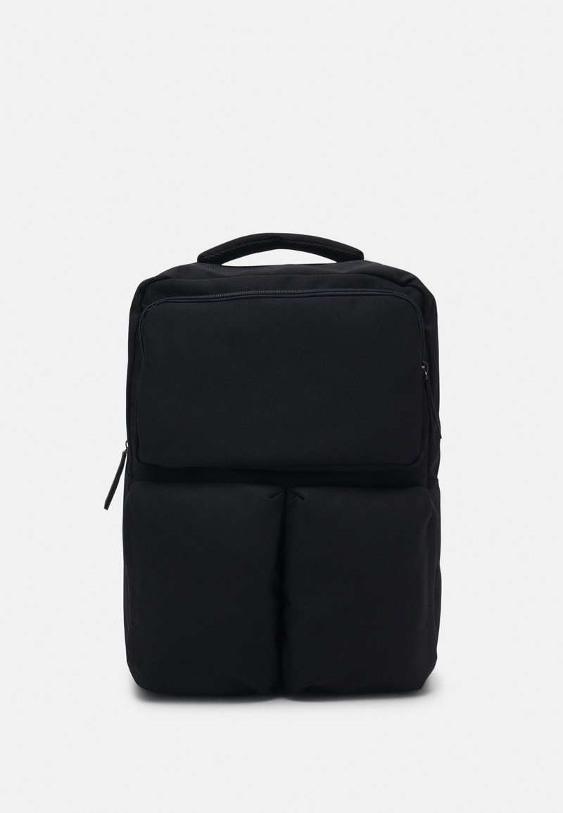 Sac à dos noir avec un devant rembourré, une poignée supérieure unique et une fermeture éclair. Présente une texture de tissu lisse et un design minimaliste.