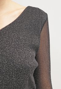 Musta toppi, jossa on teksturoitu ja kimalteleva kangas, syvällä V-päällä ja läpikuultavat pitkät hihat. Pääkankaassa on metallinhohto.