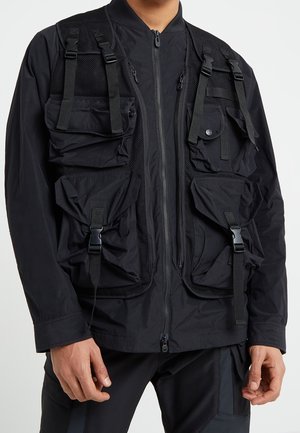 Gilet utilitaire noir avec plusieurs grandes poches sécurisées porté par-dessus une veste à fermeture éclair noire, associé à un pantalon noir.