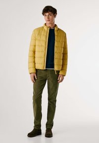 Chaqueta de plumas amarilla con patrón acolchado horizontal, cuello alto y cremallera frontal, combinada con pantalones de pana verde oscuro y botas marrones.