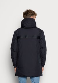 Cappotto imbottito nero con collo alto e fascia lucida con logo sulla schiena. Il design è lungo con polsini elastici e orlo dritto.