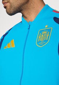 Kék atlétikai dzseki cipzárral az elején, arany spanyol nemzeti címerrel és sárga Adidas csíkokkal a vállakon.