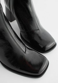 Bottines noires à finition brillante, à bout carré et au design épuré. Présente des accents de couture et un talon carré proéminent.