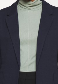 Námořnický blazer se strukturovanými rameny přes světle zelený žebrovaný rolák. Outfit má vertikální šev uprostřed vrchní části.