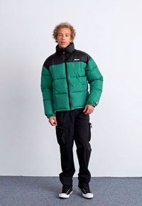 Chaqueta acolchada verde y negra con cierre delantero, que presenta un cuello alto y textura acolchada, llevada sobre pantalones cargo negros y zapatillas.