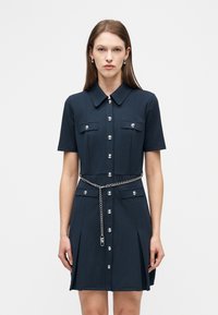 Γυναίκα που φοράει ένα σκούρο μπλε φόρεμα με κοντά μανίκια και κουμπιά, με εμπρόσθιες τσέπες και ασημένια ζώνη αλυσίδα, στέκεται μπροστά σε λευκό, απλό φόντο.