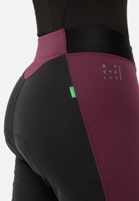 Høyt midje leggings laget av resirkulert svart og burgunder materiale, glatt tekstur og kontraststing med et grønt merke som aksent.