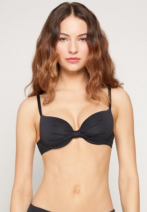 Jeune femme aux cheveux bruns ondulés portant un haut de bikini noir avec armatures et fines bretelles, debout devant un fond clair uni.