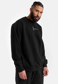 Sudadera negra de corte relajado, con puños y dobladillo acanalados, y un pequeño logo blanco en el pecho. Material de algodón, textura suave.