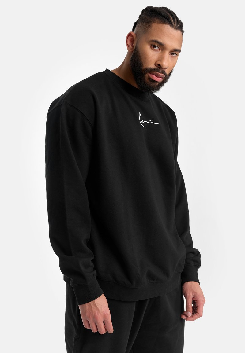 Sudadera negra de corte relajado, con puños y dobladillo acanalados, y un pequeño logo blanco en el pecho. Material de algodón, textura suave.