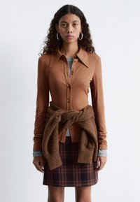 Brown long-sleeve shirt with a collar, button front, and a grey shirt underneath. A brown knitted wrap is tied around a plaid skirt.