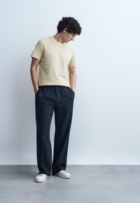 Νεαρός άνδρας με σγουρά μαλλιά που κοιτάζει προς τα κάτω, φοράει μπεζ ριγέ μπλουζάκι, μαύρο φαρδύ παντελόνι, λευκά αθλητικά παπούτσια, στέκεται μπροστά σε έναν λευκό τοίχο.