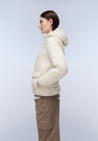 Giacca puffer crema con cappuccio, tessuto trapuntato, tasca frontale e orlo elastico. Abbinata a pantaloni cargo marroni, mostrando un profilo laterale.