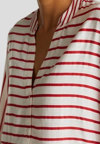 Chemise rayée en tissu léger avec des rayures horizontales alternées rouges et blanches. Elle présente un col avec fermeture par boutons.