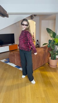 Rožinė ir juoda leopardų raštų megzta palaidinė, juodos plačios kelnės ir baltos sportbačiai. Vidaus aplinka su rudais sofais ir patalpų augalais.