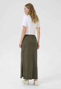 Jupe maxi vert olive avec fente latérale, taille élastique et coupe décontractée, associée à une chemise blanche à manches courtes et des baskets beiges.