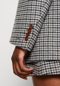 Käsi tumman ihonvärin koskettaa sinisen ja beige-houndstooth-kuvioisen bleiserin helmaa, jossa on neljä ruskeaa nappia hihassa.