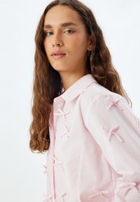 Camisa rosa claro con cuello y adornada con pequeños lazos de tela. El material parece suave con una textura lisa.