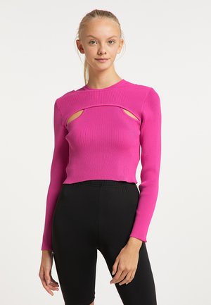 Jonge vrouw draagt een felroze geribde lange mouwen top met openingen en zwarte aangesloten broek, staand tegen een witte achtergrond.