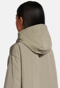 Beige Kapuzenjacke mit weicher Textur, ausgestattet mit einer gerafften Kapuze und Logodetail auf der Rückseite, entworfen für Komfort und Funktionalität.