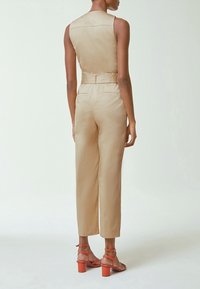 Femme portant une combinaison beige sans manches et des sandales à talons hauts orange, debout de dos sur un fond blanc uni.