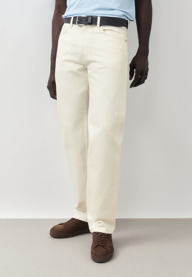 Άνδρας που φοράει εκρού παντελόνι ίσιας γραμμής, ανοιχτό μπλε πουκάμισο μέσα στην παντελόνα, μαύρη ζώνη και καφέ παπούτσια με κορδόνια, στέκεται σε γκρι πάτωμα.