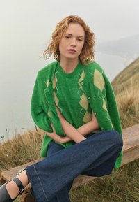 Groene wollen cardigan met diamantpatronen in geel en een bijpassende groene trui eronder, gecombineerd met donkere denim wijde broek en zwarte schoenen.