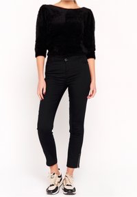 Mujer vistiendo una blusa negra de manga tres cuartos de felpa, pantalones skinny negros de talle alto con cremalleras en los tobillos y zapatillas beige y negras.