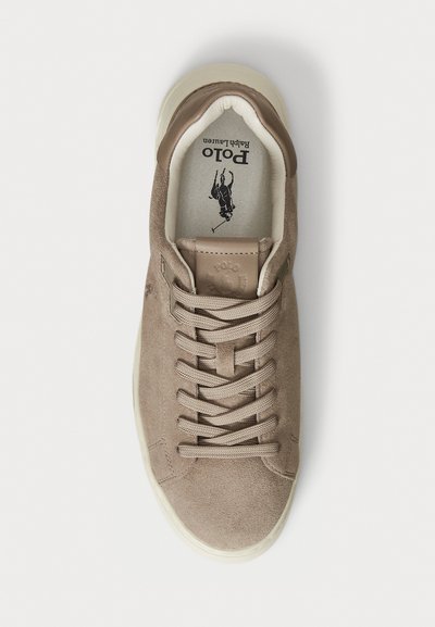 Sneaker bej din suede cu vârf rotund, șireturi plate și un logo pe limbă. Talpa este de culoare crem, cu o textură netedă.