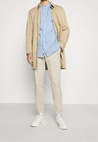 Un trench-coat beige, une chemise bleu clair à boutons, un pantalon crème et des baskets blanches. Un design simple avec un look décontracté et des tissus texturés.