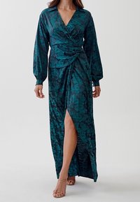 Lång tealblå blommig omlotklänning med långa ärmar, V-öppning och sprund upp till låret. Tillverkad av ett mjukt, flytande tyg med en behaglig textur.
