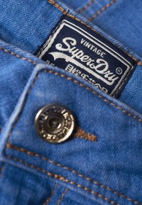 Tela de mezclilla en azul claro, con un botón de metal y una etiqueta tejida con "SuperDry" en blanco sobre un fondo azul marino.