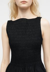 Γυναίκα που φοράει μαύρο φόρεμα χωρίς μανίκια με ραμμένη μπούστο και στρογγυλή λαιμόκοψη, που αφήνει ακάλυπτους τους ώμους και το άνω μέρος του στήθους, με ελαφρώς κυματιστά ανοιχτά μαλλιά.