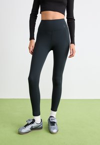 Person som bär svarta högmidjade leggings, vita strumpor och silverfärgade Nike-sneakers står på ett grönt golv framför en vit bakgrund.