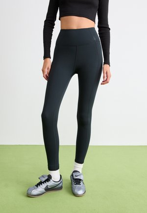 Person iført sorte højtaljede leggings, hvide sokker og sølvfarvede Nike sneakers, stående på et grønt gulv foran en hvid baggrund.