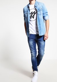 Světle modrá denimová košile přes bílý tričko s grafikou, spárována se středně modrými džíny a bílými teniskami. Jednoduchý design s uvolněným střihem.