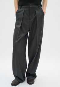 Pantalones gris con plegados y corte a medida, confeccionados con un tejido texturizado; presentan pasadores para cinturón y bolsillos laterales, combinados con zapatos marrón oscuro.