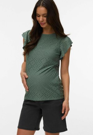 Zwangere vrouw met lang donker haar, gekleed in een groene blouse met oogjes en zwarte korte broek, die haar buik zachtjes vasthoudt en naar beneden kijkt.